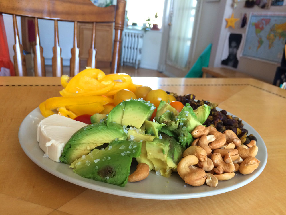 plate on a kitchen table with fresh cheese, sliced yellow peppers and avocado, cashews, sweet grape tomatoes, and black beans